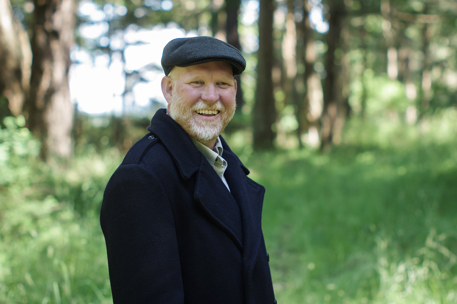 Photograph of a man in his late forties wearing a wool Thread Theory peacoat and a flat cap, smiling and looking at the camera. He's standing in front of trees and grass that are out of focus in the background.