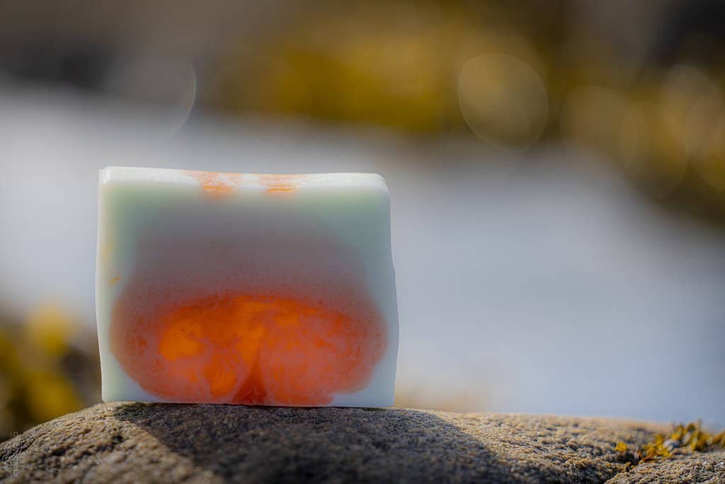 Close up of a bar of UasaU soap sitting on a rock. The soap is glowing as it is back lit by the sun. The soap is a mottled orange and white.