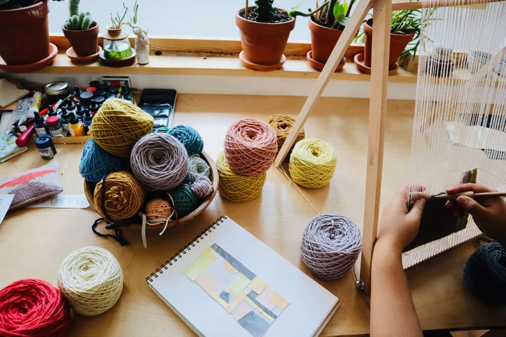 Still life photograph of spools of different coloured spools of yarn, spilling out on a table. Arounna's hands are pictured working on her loon and the reference artwork is resting on the table in front of her.