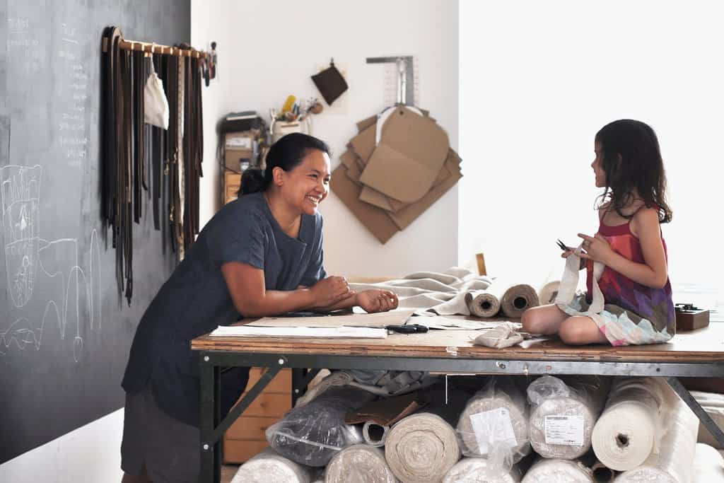 Photograph of Arounna leaning on her work table looking at her young daughter who is sitting on top of the table. They are smiling at each other. There are several roles of fabric tucked under the table.