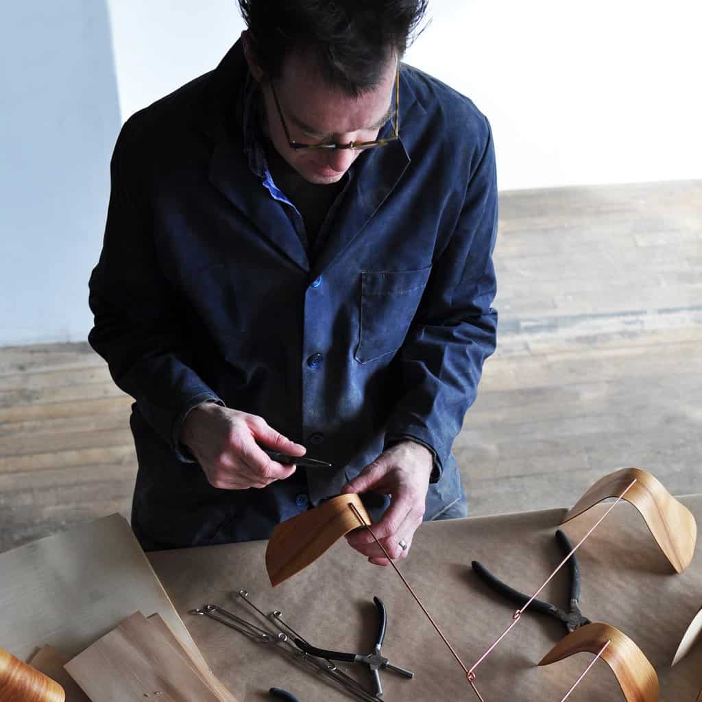 Photograph from slightly overhead of John hand making his wooden mobile. He is wearing a dark denim button down shirt.