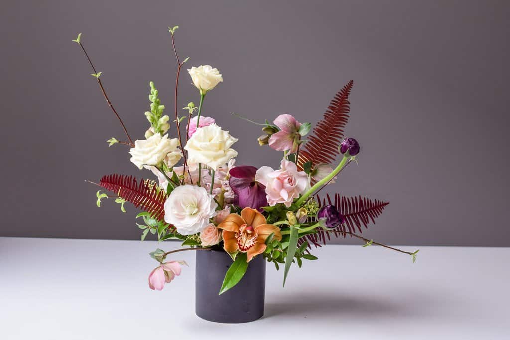A gray vase filled with a flower bouquet sits on a surface against a plain backdrop