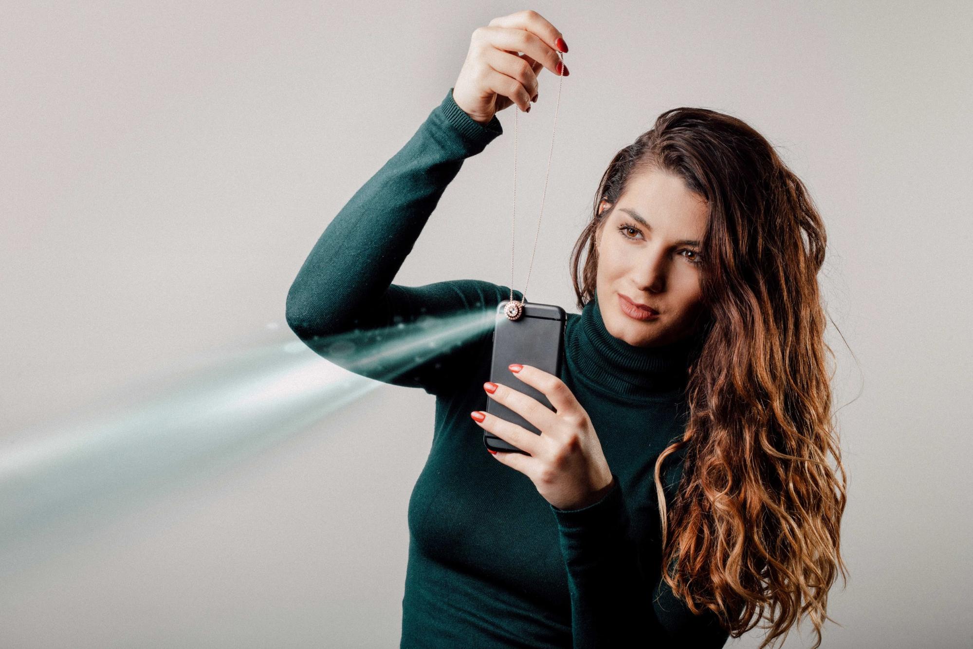 A model in a green sweater is holding a Revival Jewelry locket while shining a light from her mobile phone to project an image from the gem of the pendant.