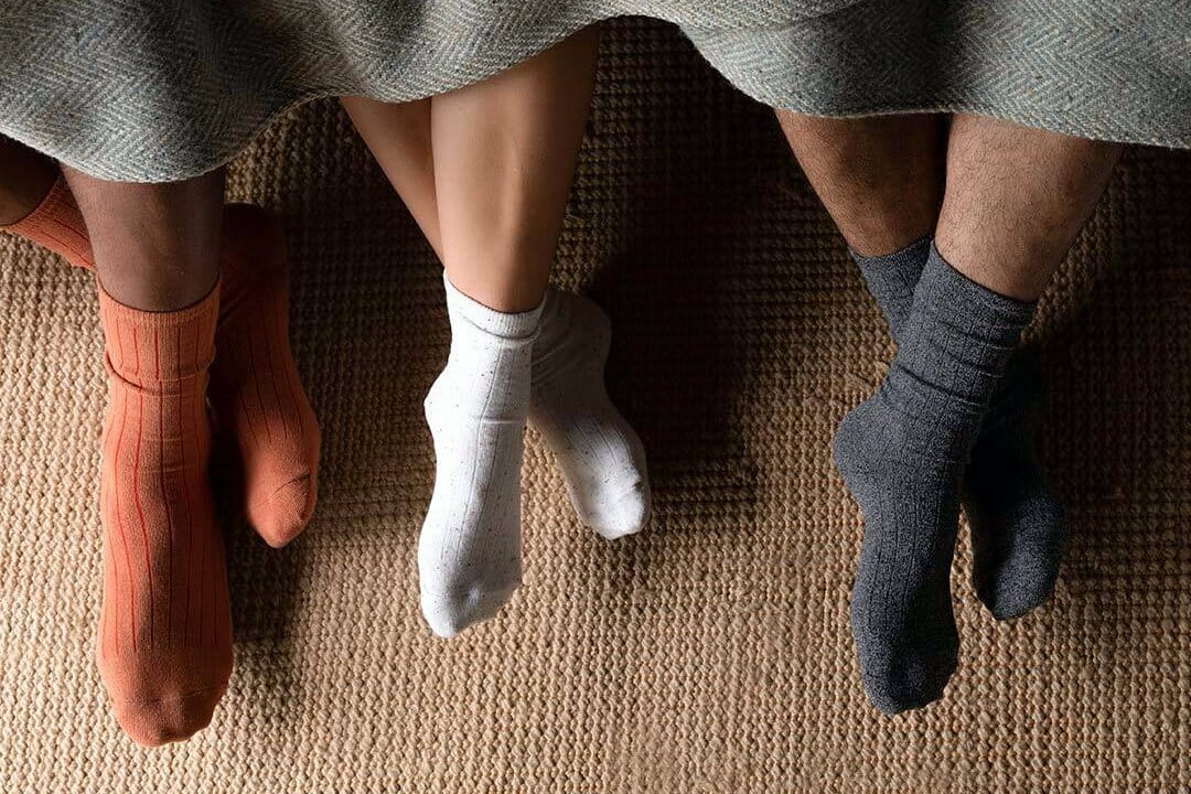Three pairs of legs poke out from under a blanket, all wearing cozy socks