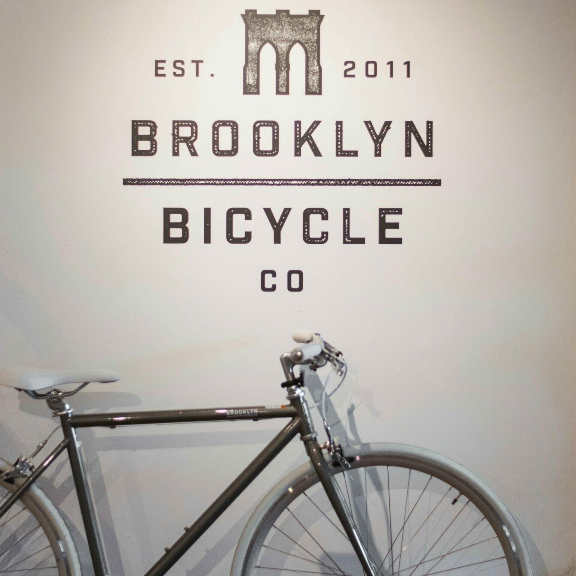 A grey bicycle by Brooklyn Bicycle Co. in their showroom against a wall with their logo.