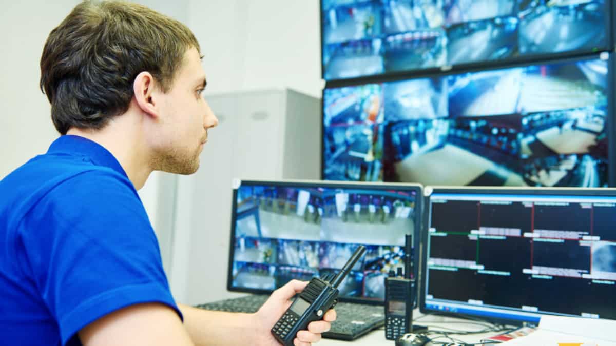 A man in a blue shirt is benefiting from a CMMS as he looks at several monitors.