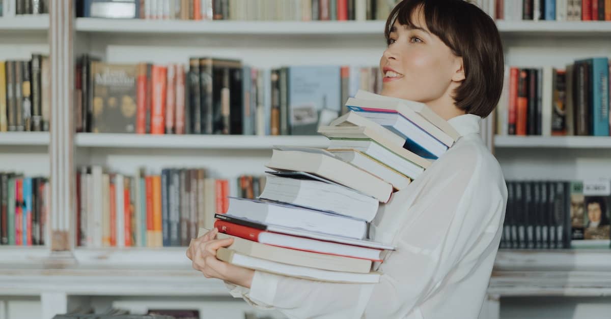 A woman holding a stack of books in front of a bookshelf, showcasing her love for literature.