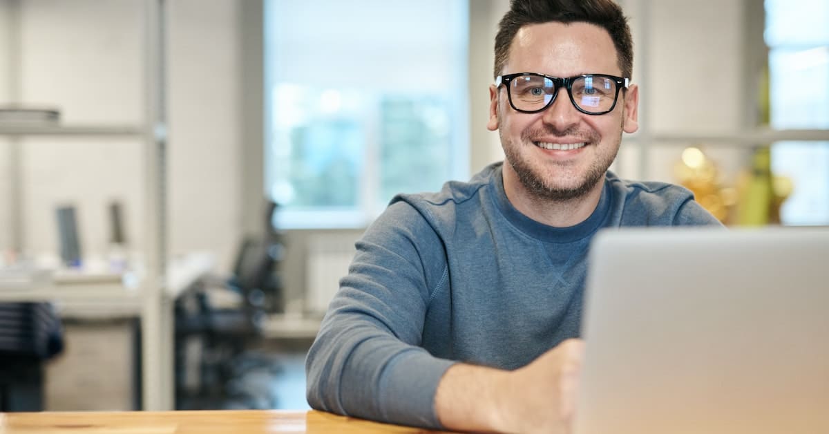 A man wearing glasses is smiling while using a laptop in a business setting.