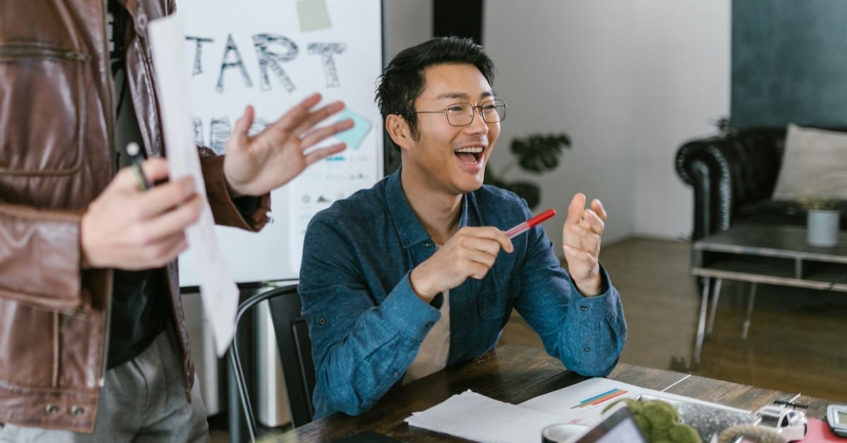 Two Asian men laughing at a table in an office, showcasing the traits of an entrepreneurship mindset.