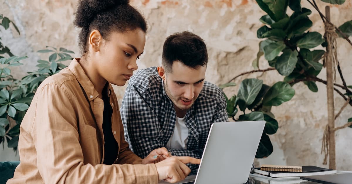 Two people sitting at a table, viewing a laptop while discussing potential digital marketing strategies.