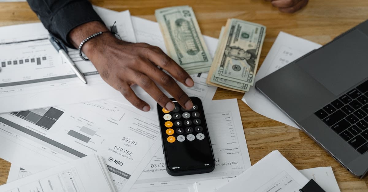 A man is sitting at a desk with a laptop and money, holding a remote control.