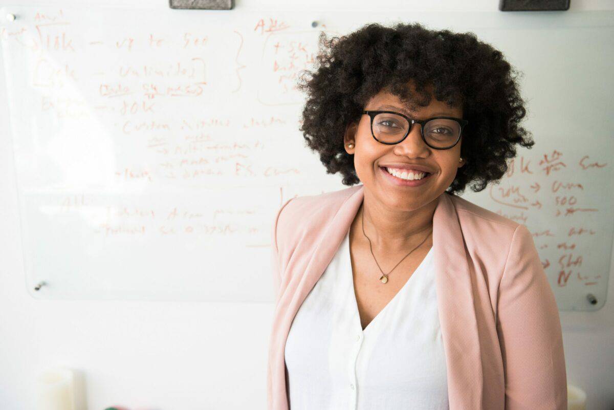 A cheerful woman with curly hair wearing glasses and a pink blazer stands in front of a whiteboard with mathematical equations, embodying the entrepreneurial fire in tech.