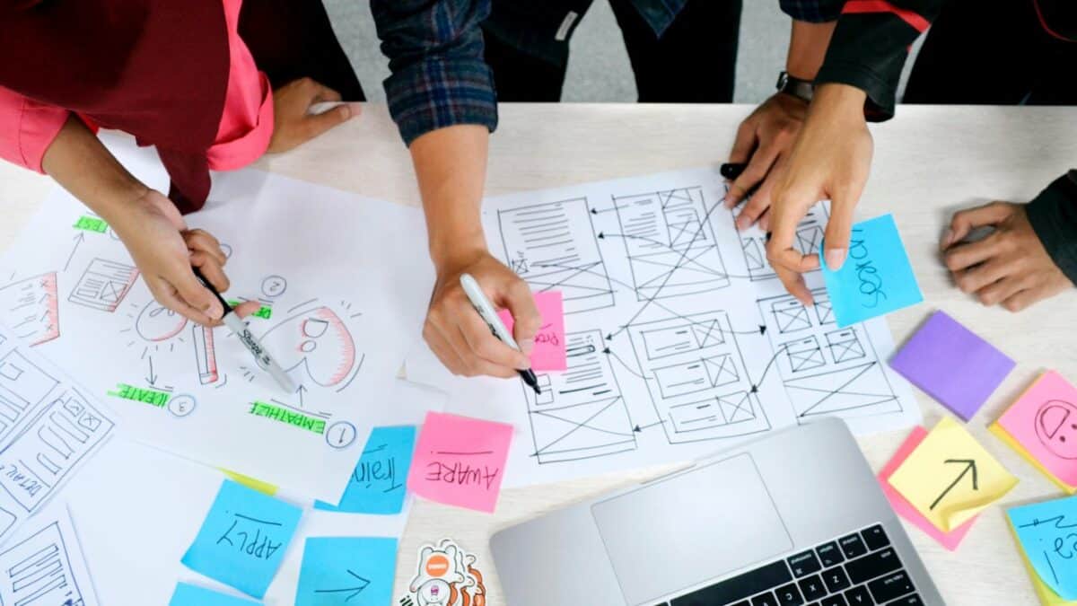 Top view of a collaborative IT outsourcing workspace with hands of multiple people organizing sticky notes and sketches on paper, with a laptop nearby.