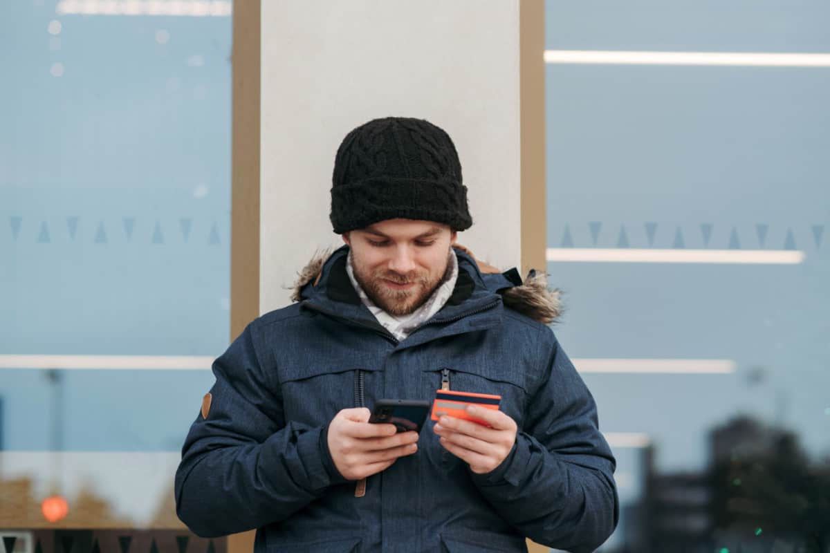 A man in winter clothing using a smartphone optimized for SEO while holding a credit card outside a store.