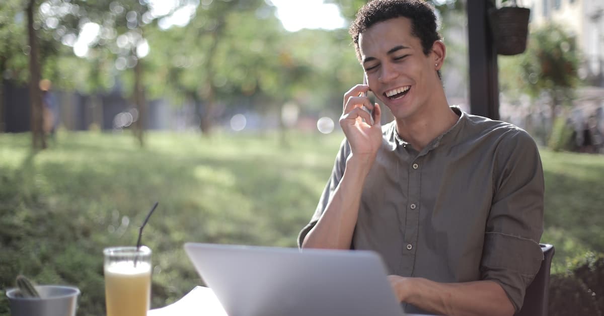 Un joven ríe mientras habla por su teléfono inteligente, sentado en una mesa de café al aire libre con una computadora portátil y un vaso de jugo, discutiendo sobre ética del marketing.