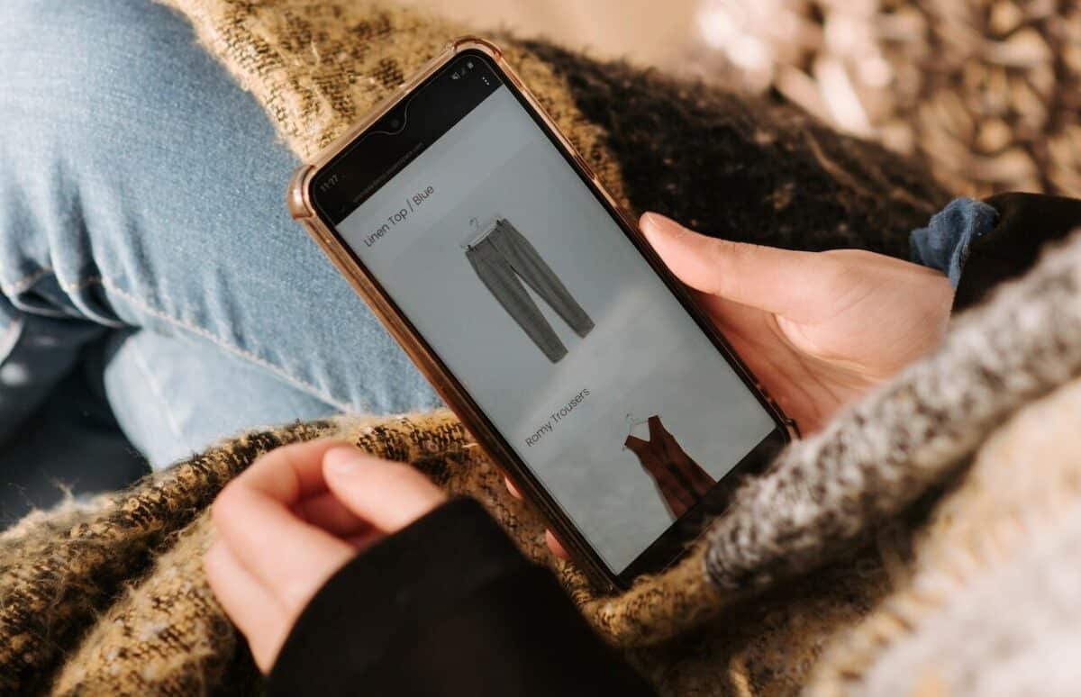 A person holding a smartphone displaying a D2C ecommerce shopping page with images of pants and a top, wrapped in a cozy blanket.