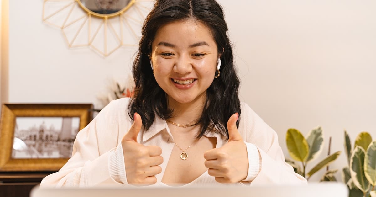 A young woman wearing headphones sits at a desk, smiling and giving two thumbs up in a well-lit room with decorative plants and a photo frame in the background, possibly brainstorming website marketing strategies.