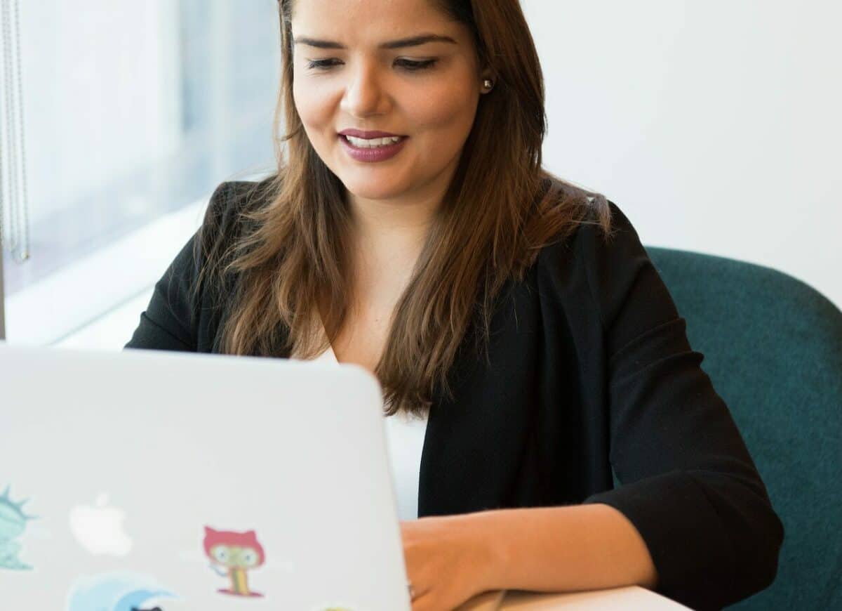 A woman in a black blazer smiling while using a laptop covered with colorful stickers in a modern office setting, engaged in e-commerce.