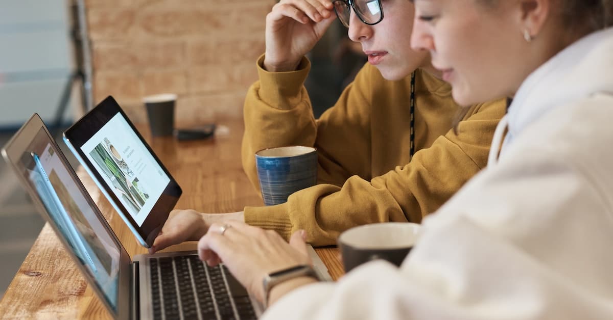 Two women conducting market research by examining a laptop screen together at a wooden table with coffee cups visible; one is typing and the other is pointing at the screen.