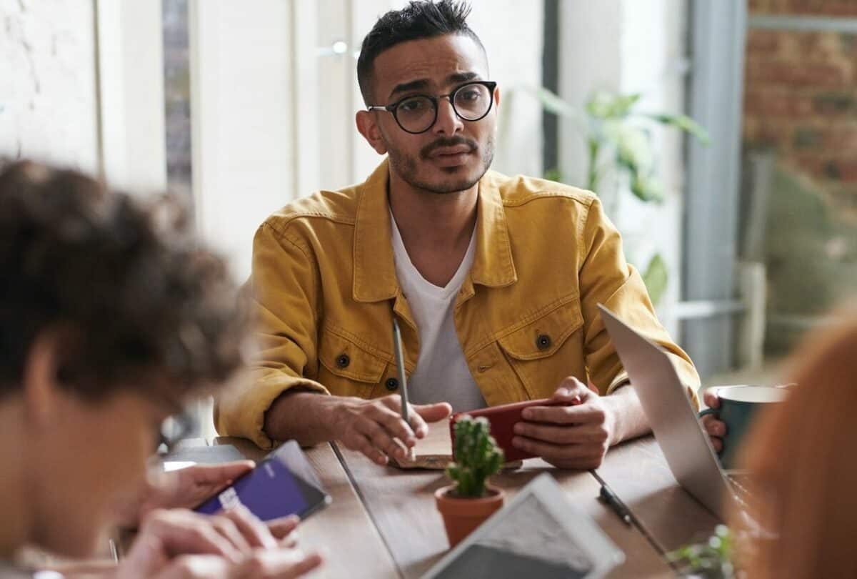 A man in a yellow jacket and glasses sits at a table with others, discussing best practices and looking attentive while holding a smartphone.