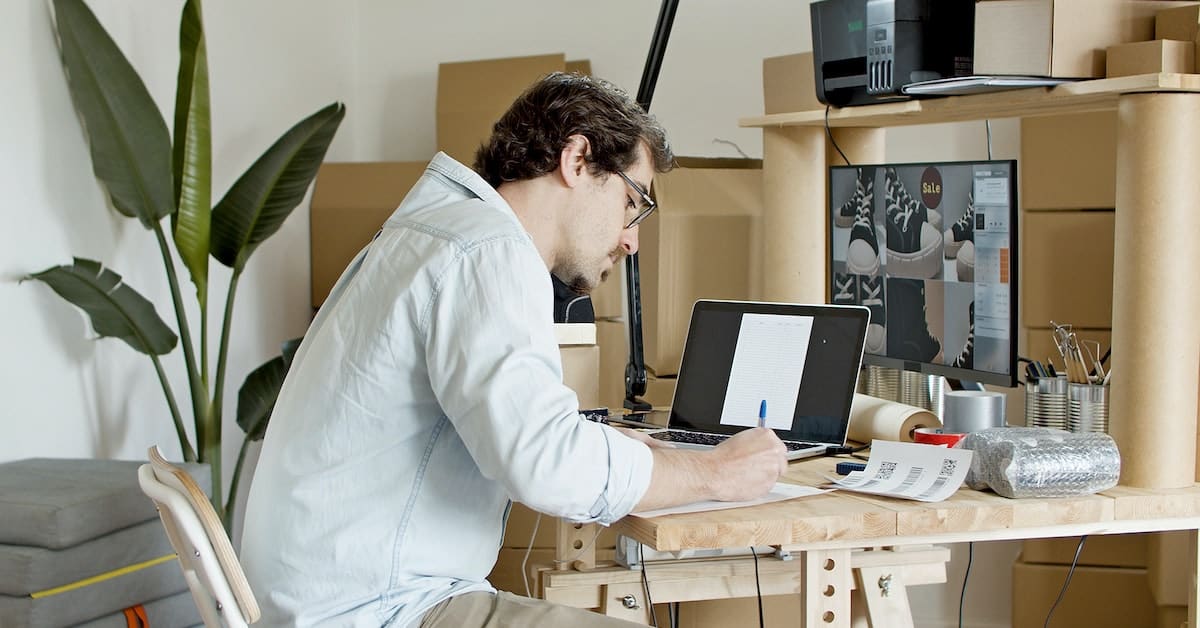 A man wearing glasses and a white shirt working on SEO keywords for an online store at a cluttered desk in an office with plants and shelves.