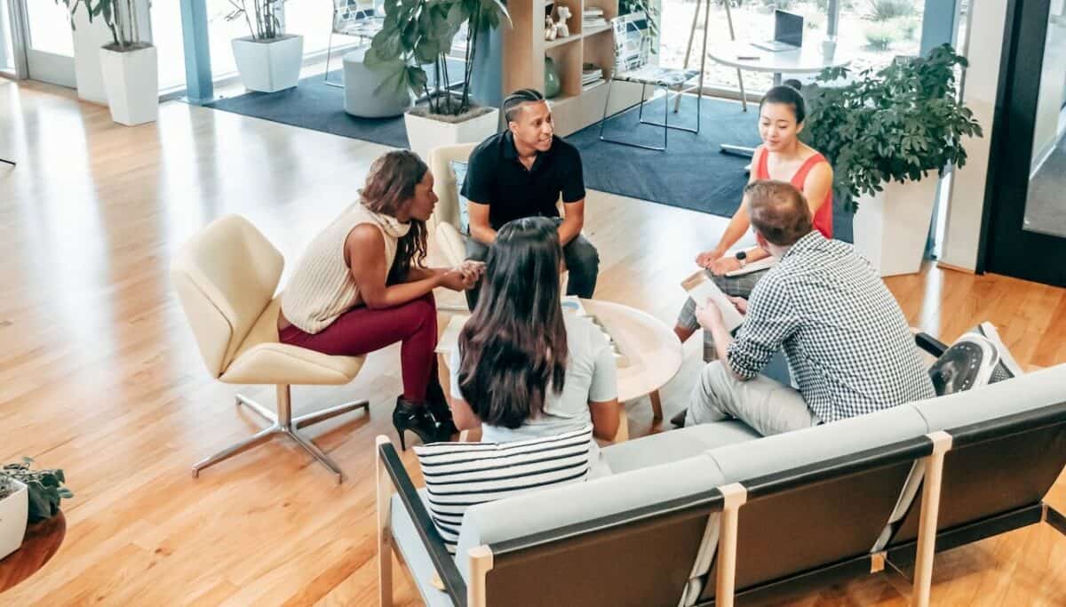 A diverse group of five professionals engaged in a discussion about the best marketing strategies, sitting around a low table in a modern office lounge area.