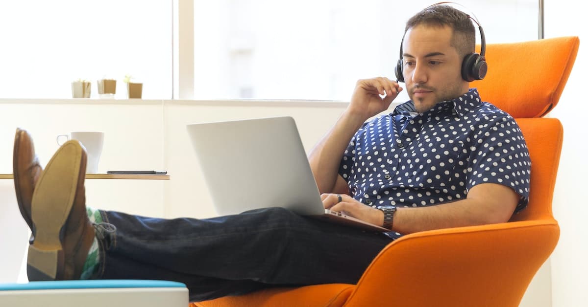 A man wearing headphones uses a laptop while sitting with his feet up in an orange chair in a modern office setting, focusing on web development services.