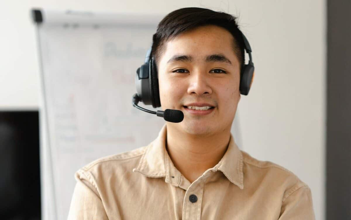A young Asian man wearing a headset with a microphone, smiling at the camera in an IT management office setting.