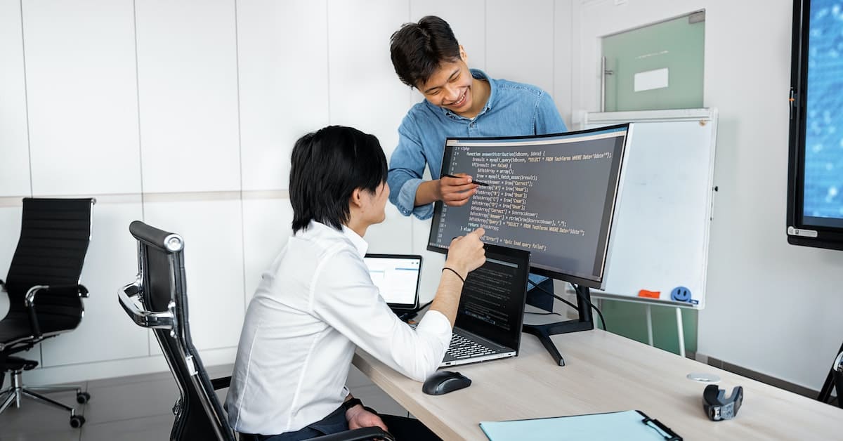 Two professionals collaborating on offshore software development at a desk, focusing on a laptop and monitor displaying code in a modern office.