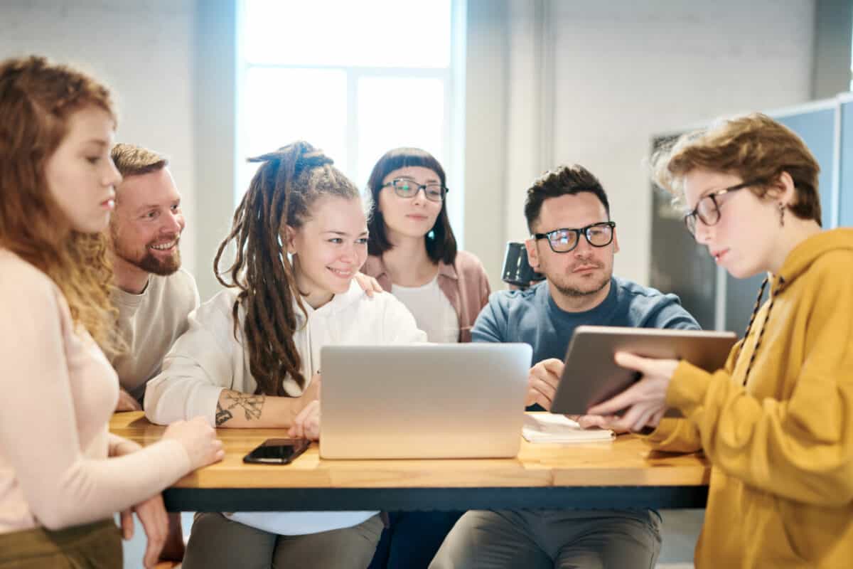 A diverse group of six young professionals from boutique advertising agencies engaged in a discussion around a laptop in a bright office setting.