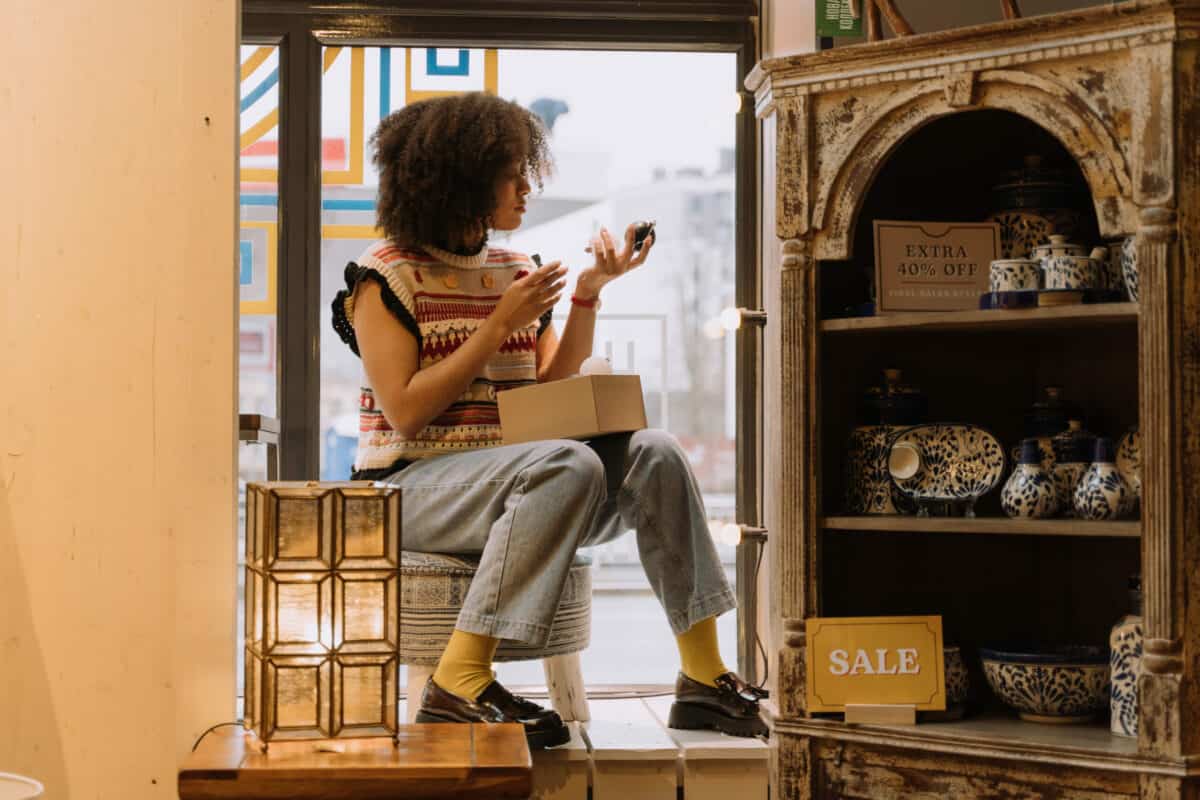 A young woman sits by a store window, looking at her phone, with boxes and a promotions sign beside her.