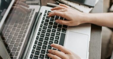 A person typing on a laptop keyboard, with another laptop and a smartphone visible on the desk, working on a public relations project for Glazers Media.