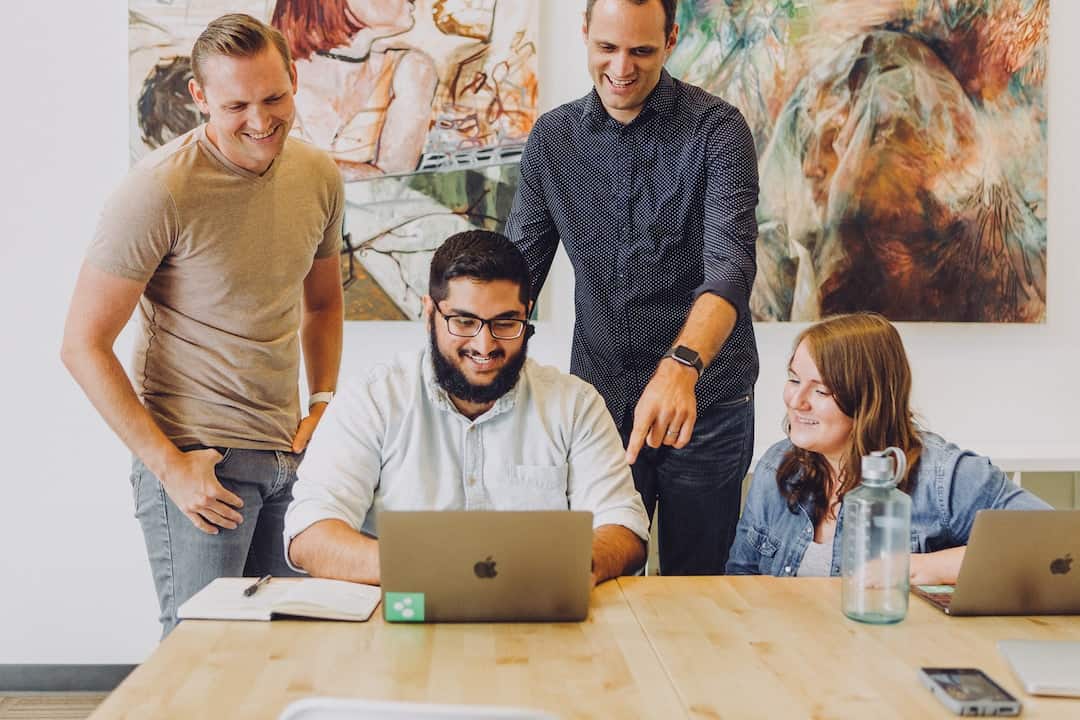 Four colleagues laugh and discuss operational reporting solutions around a laptop in a modern office; artwork hangs on the wall behind them.