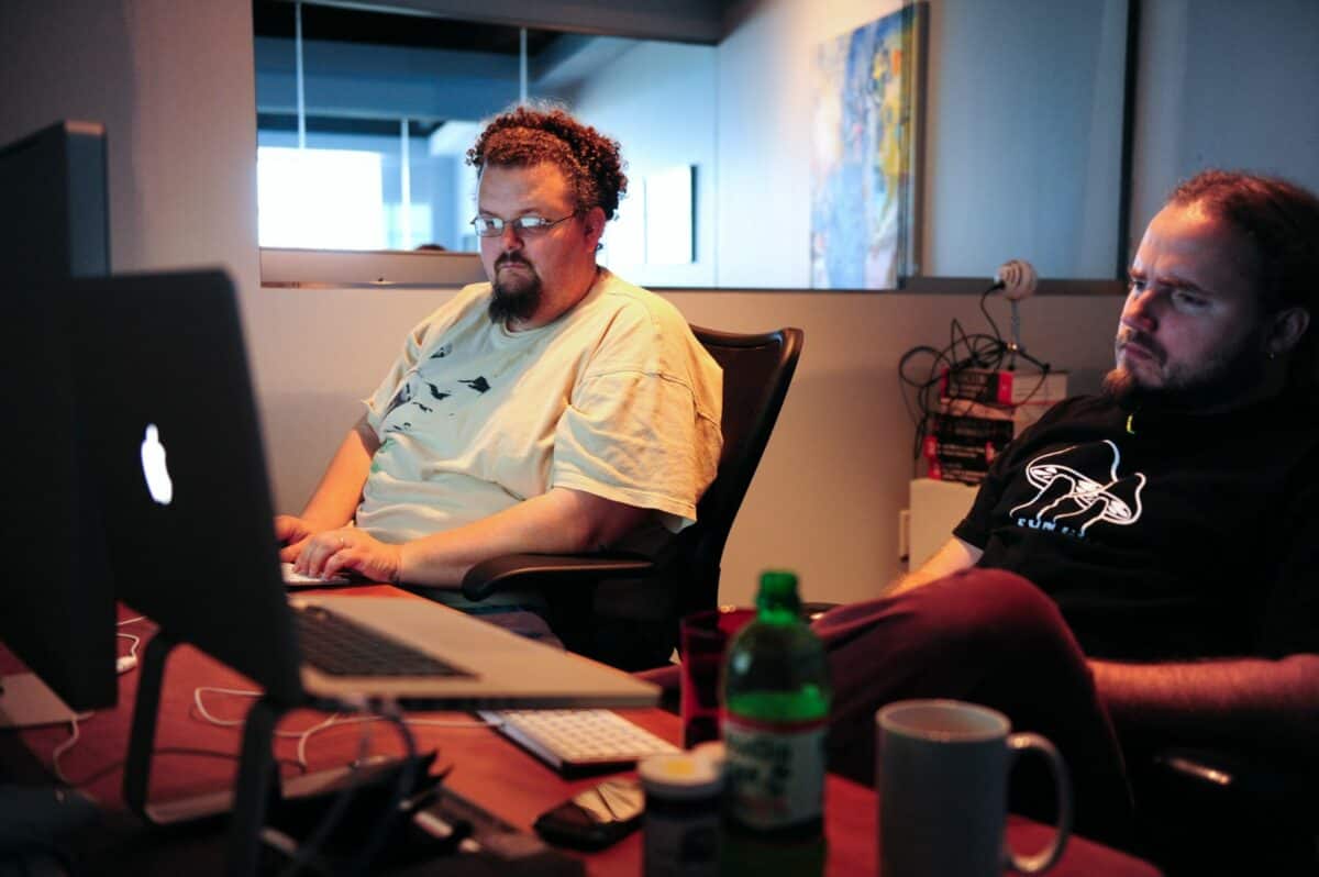Two men focusing on an AWS Certified Solutions Architect exam preparation on a computer screen in a dimly lit office with posters on the wall and drinks on the desk.