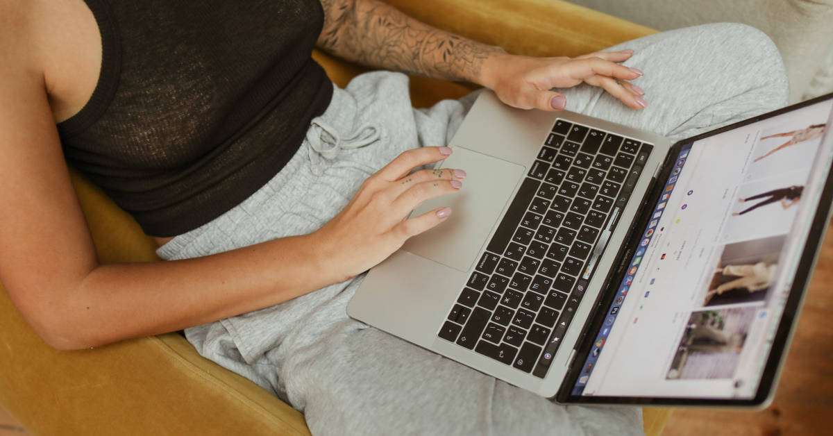 A person lounging on a chair, using a laptop for ecommerce marketing, dressed in casual grey sweatpants and a black top, displaying a tattoo on their arm.