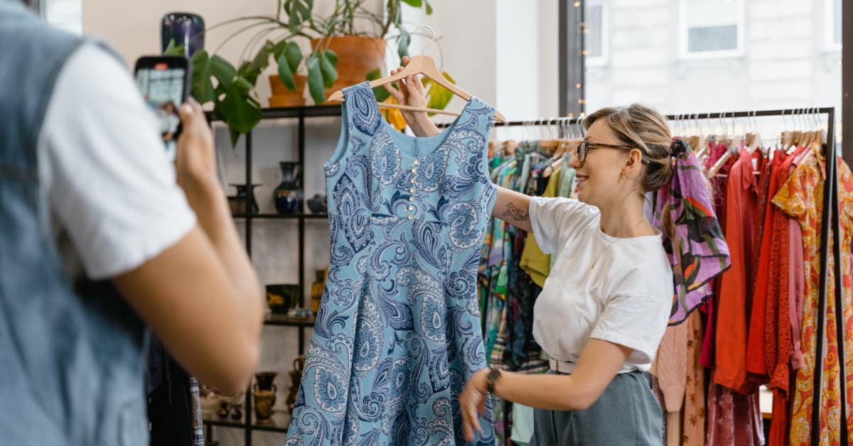 A woman in a clothing store holds up a blue patterned dress, smiling at someone taking her photo for an ecommerce marketing campaign, surrounded by various colorful garments.