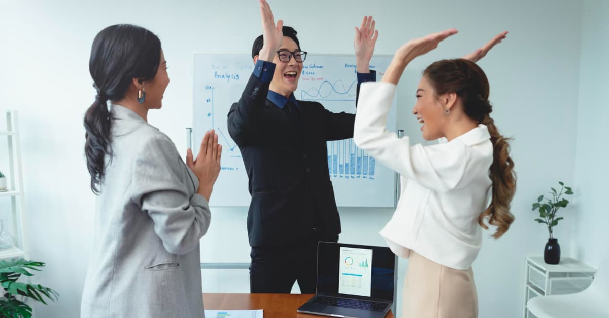 Three people in business attire celebrate in an office, standing around a desk with a laptop. A whiteboard with charts is visible in the background, illustrating their recent success in boosting customer engagement.