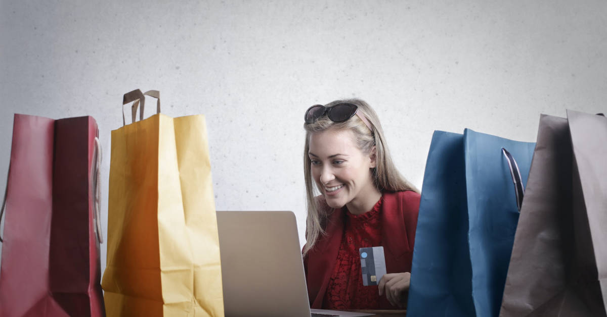 A woman smiling while shopping online with a laptop, surrounded by colorful shopping bags made of eco-packaging and holding a credit card.