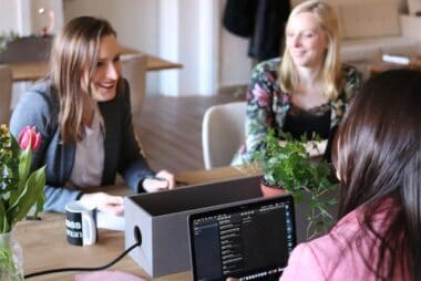 Three women discussing over a laptop at an e-commerce helpdesk meeting, with coffee mugs and flowers on the table.