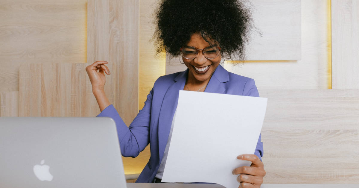 A joyful black woman in a purple blazer, wearing glasses, reads a document about e-commerce success and celebrates at her desk with a laptop.