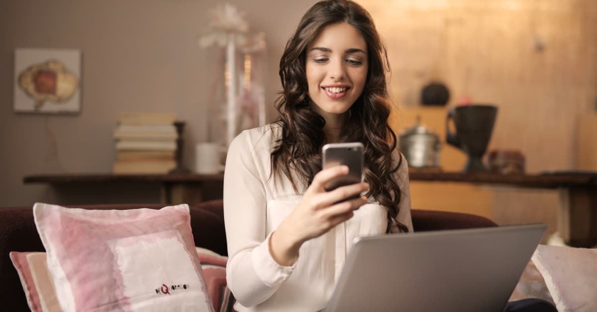A woman smiles while using her smartphone to collaborate with micro-influencers and laptop in a cozy living room setting.
