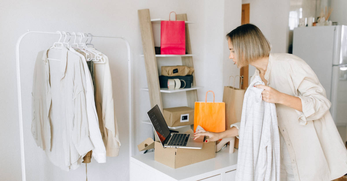 Woman shopping online for luxury brands, choosing clothes with a laptop, box, and shopping bags in a bright room.