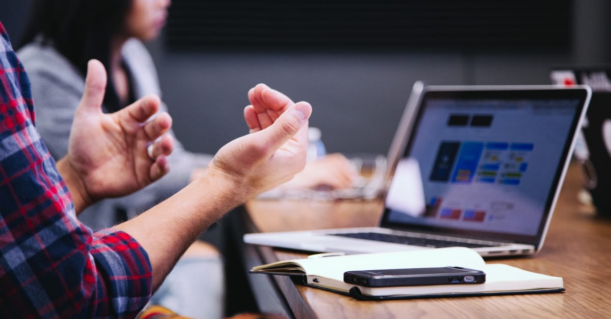 A person in a plaid shirt gestures while discussing an e-commerce project in a meeting with a laptop displaying graphs and a smartphone on the table.