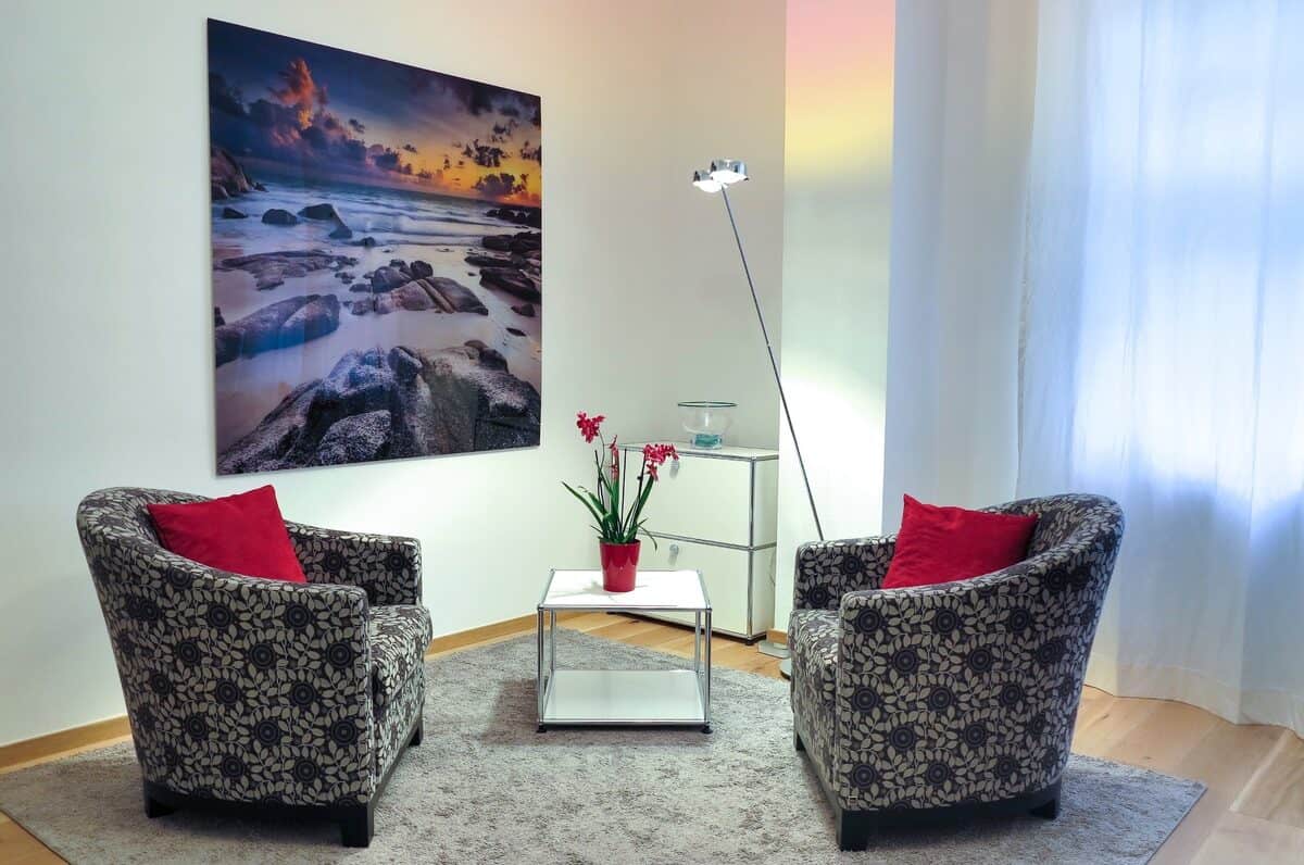 Two eco-friendly armchairs with floral patterns, red cushions, and a small table with a flower pot are arranged in a cozy corner. A large framed beach photo and a standing lamp are in the background, creating an inviting space that champions sustainability.