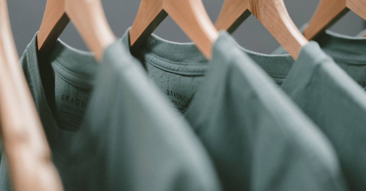 A close-up of green t-shirts from a clothing brand hanging neatly on wooden hangers against a soft focus background.