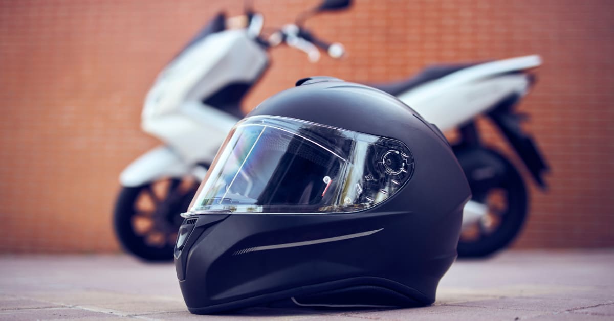Black motorcycle helmet on the ground in front of a white scooter against a brick wall background, serving as the perfect visual for an ultimate guide to choosing the right motorcycle helmet.