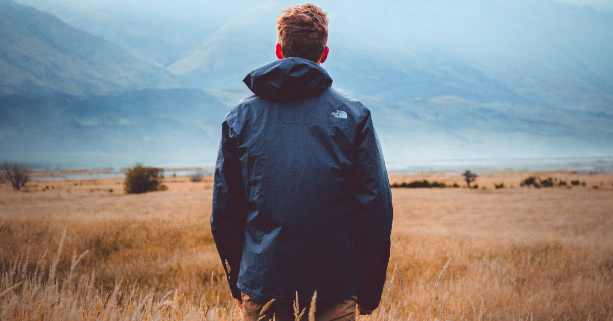 Man in a black jacket standing in a field with distant mountains and overcast sky, ideal for brandcasting.