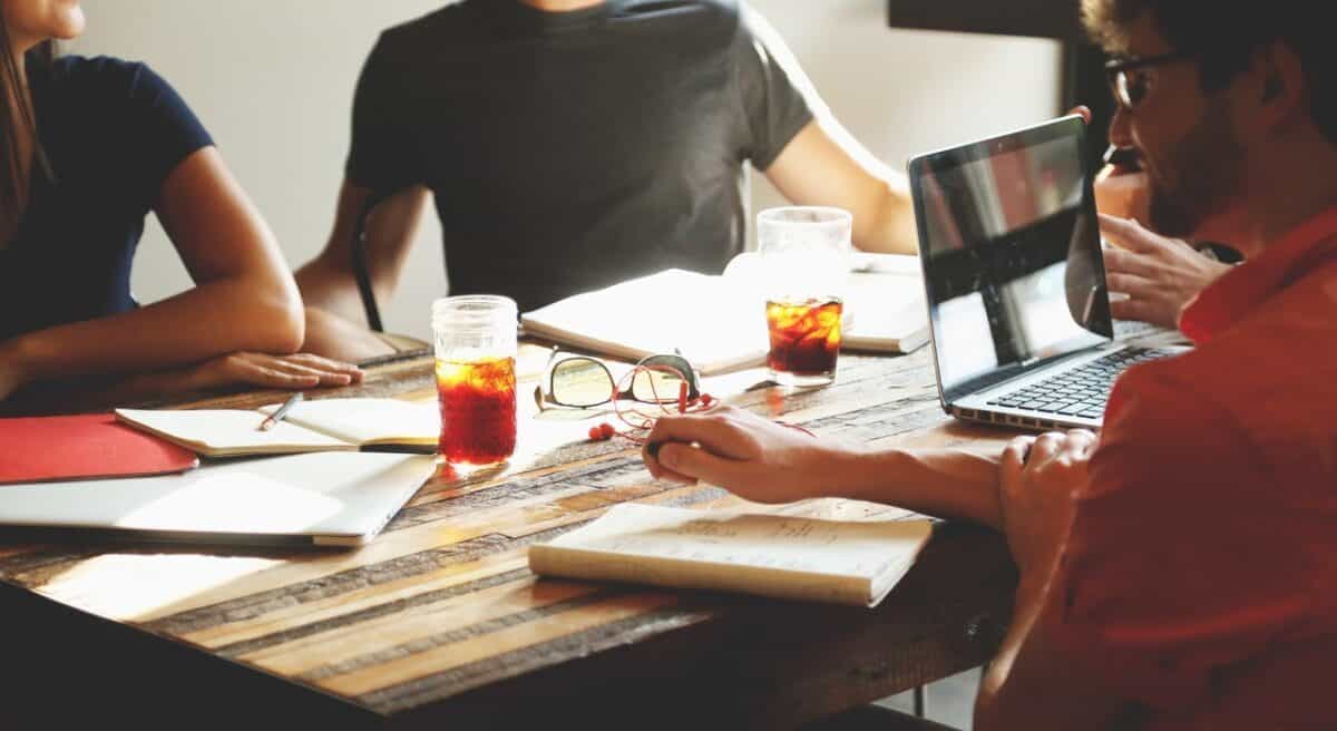 Three people in a meeting, seated around a wooden table with notebooks, laptops, and drinks. Sunlight streams in, creating a bright atmosphere as they discuss the future of innovation and explore choosing innovation software to enhance their workflow.