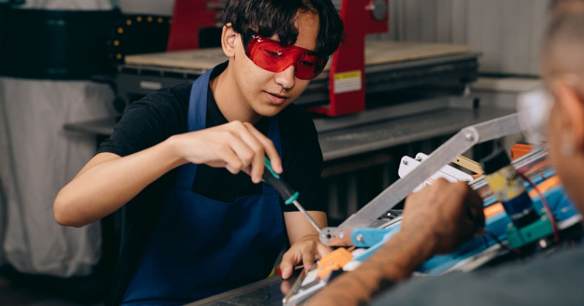 Young Asian man in safety goggles and apron using a screwdriver on a mechanical part in a sustainable manufacturing workshop, interacting with a colleague.