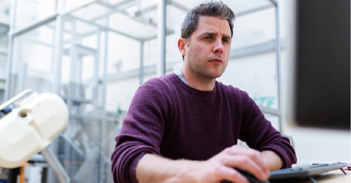 A man in a purple sweater sitting at a desk in an office, looking focused while managing online reputation on a laptop.
